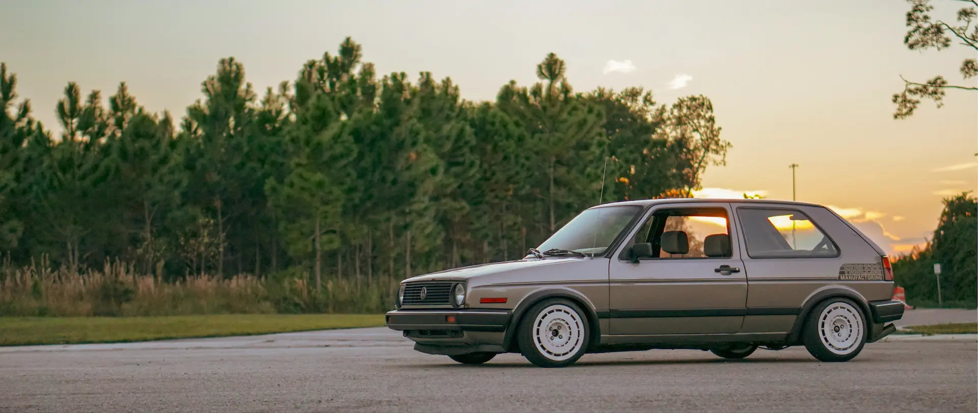 A classic Volkswagen Golf Mk II in metallic taupe is parked on an asphalt road with a forest and sunset in the background.